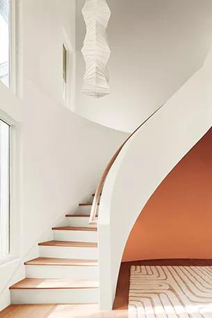 Una escalera curva pintada de blanco impuro con una audaz pared de acento roja debajo de ella, una alfombra de área neutra, una brillante luz natural que entra a través de grandes ventanas y una lámpara de papel de arroz