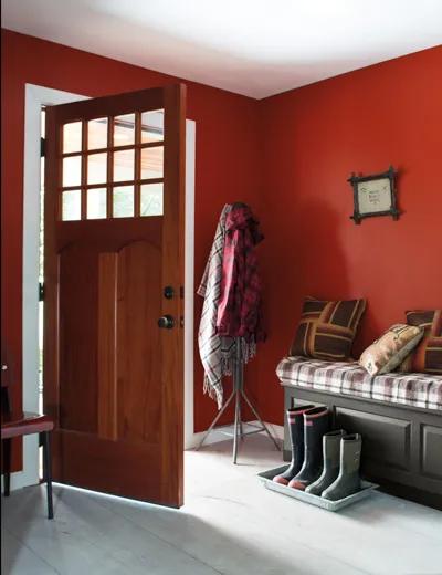 Una entrada de casa pintada de rojo con un banco gris oscuro y una puerta de madera abierta.