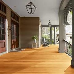 Un espacioso porche delantero cubierto con revestimientos exteriores pintados de color beige, columnas de color verde apagado y techo blanco, que cuenta con hermosas puertas principales y piso teñido con Woodluxe® Tinte para exteriores Traslúcido en Natural ES-10.