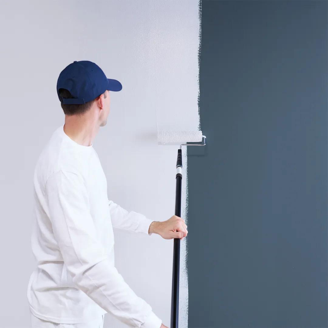 Un hombre aplicando imprimador con un rodillo sobre una pared pintada de azul oscuro.