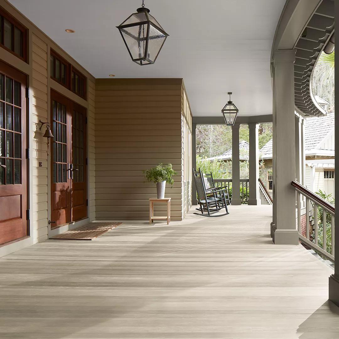 Un espacioso porche delantero cubierto con revestimientos exteriores pintados de color Beige, columnas de color Verde apagado y techo Blanco, cuenta con un hermoso piso y puertas principales con Woodluxe® Tinte para exteriores traslúcido en Gris Blanquecino ES-78.