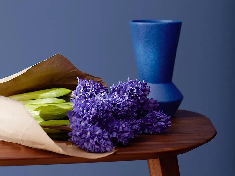 Una pared de azul profundo con marco decorativo detrás de una pequeña mesa de madera que sostiene un jarrón de cobalto y un ramo de flores púrpuras envuelto en papel marrón.