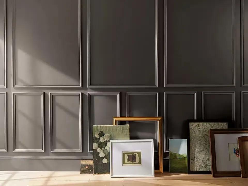 Obras de arte y marcos dispuestos en el piso contra una pared pintada de marrón chocolate oscuro con molduras decorativas.