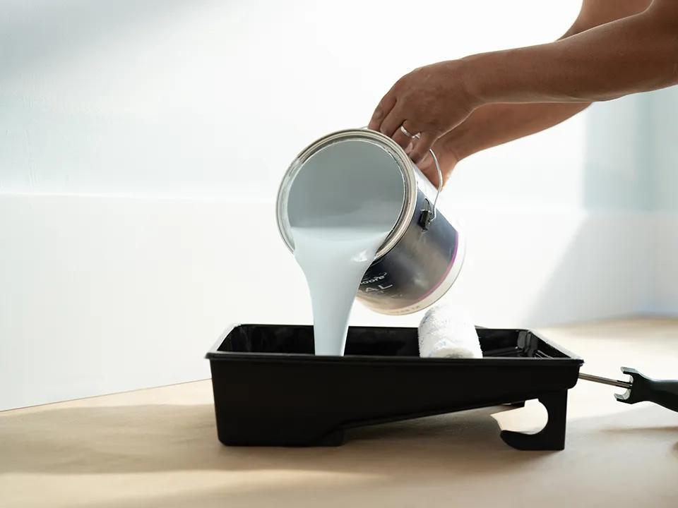 Una persona vierte una lata de pintura para interiores Regal® Select en color Estanque Tranquilo en una cubeta de pintura, cerca de una pared blanca.