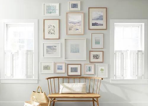 Una pared de pasillo pintada de blanco impuro con dos ventanas con postigos blancas y una colección de arte enmarcado arriba de un banco de madera.