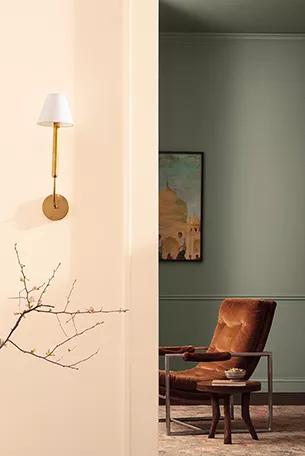 Un pasillo pintado en rosado y blanco impuro con una lámpara de pared de latón y ramas decorativas, y una entrada a una sala de estar con una pared pintada de verde oscuro, una silla tapizada en rojo y una mesa auxiliar redonda de madera.