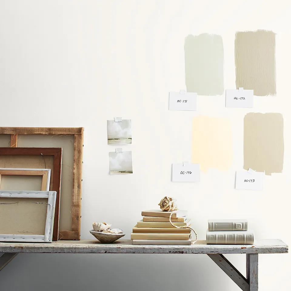Cuatro muestras de colores pintadas con números de color encintados sobre una pared blanca arriba de una mesa con libros y lienzos apoyados.