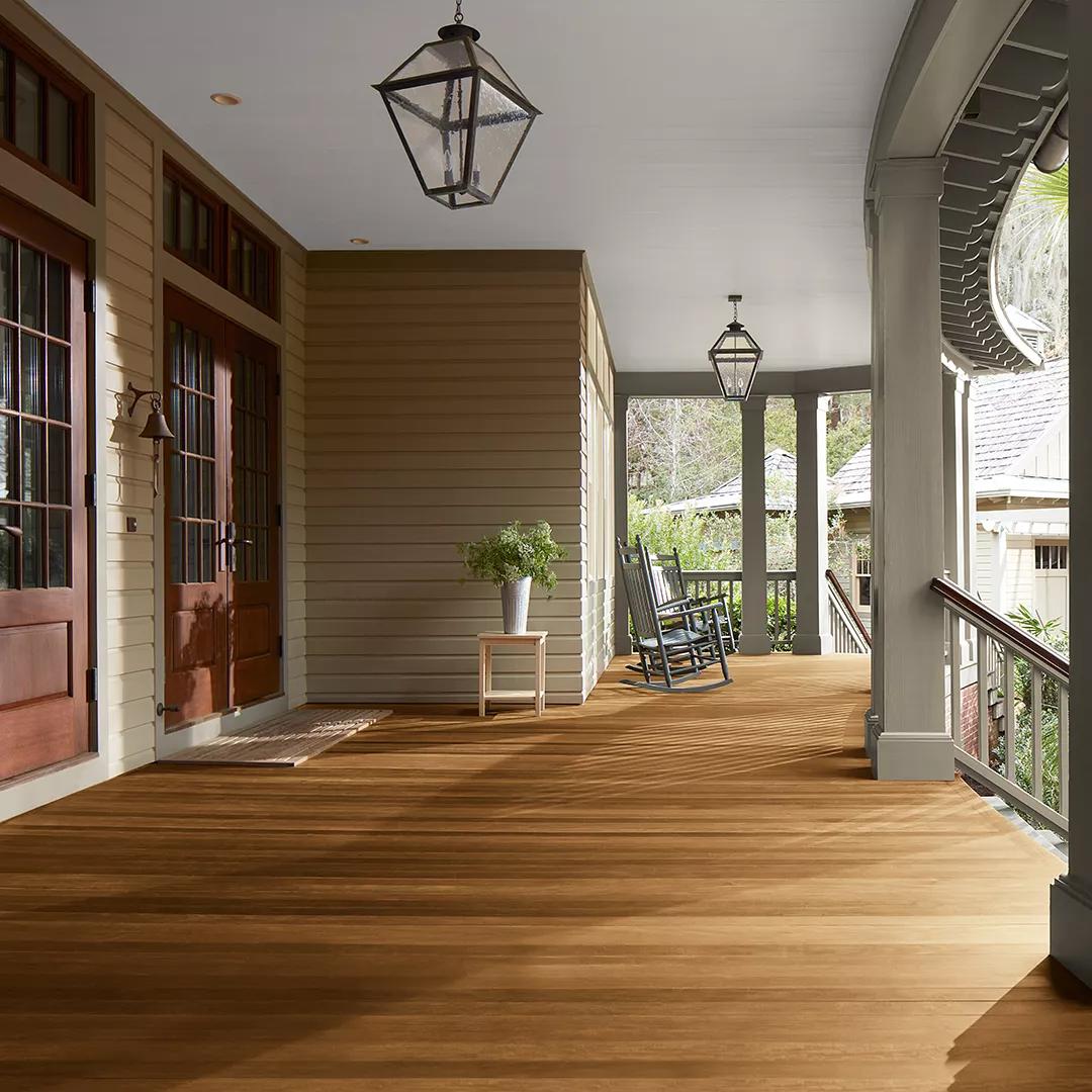 Un espacioso porche delantero cubierto con revestimientos exteriores pintados de color Beige, columnas de color Verde apagado y techo Blanco, que cuenta con hermosas puertas principales y piso teñido con Woodluxe® Tinte para exteriores Traslúcido en Marrón Castaña ES-65.