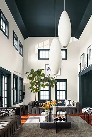 Una gran sala dramática con un techo alto y marco para ventanas pintados de Verde Intenso, paredes pintadas en blanco impuro, grandes sofás de color gris carboncillo, una mesa de cóctel lacada en negro y accesorios de iluminación colgantes en forma de óvalo de gran tamaño.