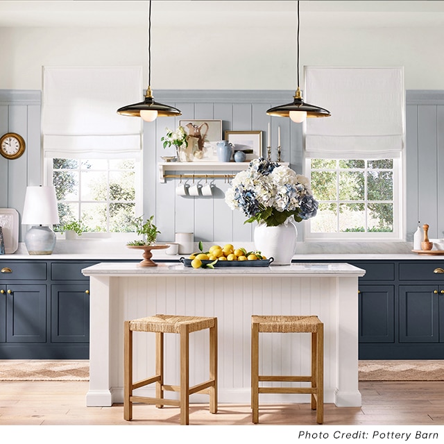 Una cocina con paredes de madera pintadas de azul en la sección inferior de las paredes, parte superior pintada de color blanco impuro, gabinetes pintados de azul, una isla pintada de blanco y varias decoraciones en encimeras de mármol.